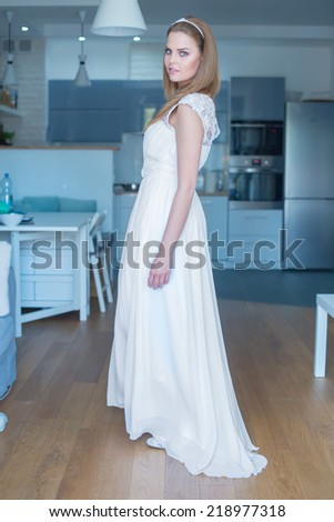 Woman Wearing White Wedding Dress Standing in Kitchen and Looking Over Shoulder at Camera