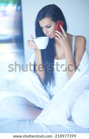 Young woman drinking coffee as she talks on her mobile phone while relaxing in bed on a sunny morning
