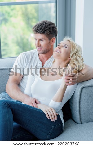 Young couple spending a relaxing day at home reclining together on a sofa below a window overlooking the garden looking off to the side with a smile