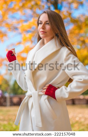 Elegant confident young woman in a stylish white overcoat and red mittens standing looking into the distance in a colorful autumn park with yellow foliage