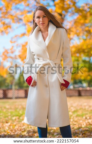 Fashionable confident young woman wearing a stylish white overcoat and red mittens standing outdoors in a colorful autumn park looking at the camera