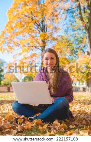 Pretty friendly woman sitting cross-legged on the ground amongst colorful leaves in an autumn park with her laptop on her lap