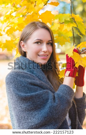 Smiling woman gathering autumn leaves in her hands wearing red mittens posing against bright yellow fall foliage