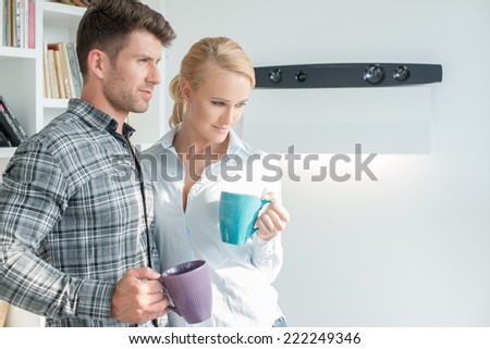 Young couple standing drinking coffee in their living room standing side by side looking off to the right of the frame