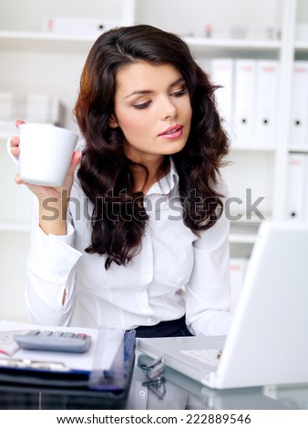 Young businesswoman with long wavy brown hair working on her laptop and drinking a mug of coffee as she sits at her desk in the office