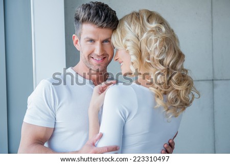 Handsome young man with unshaven stubble embracing his blond wife and smiling over her shoulder at the camera
