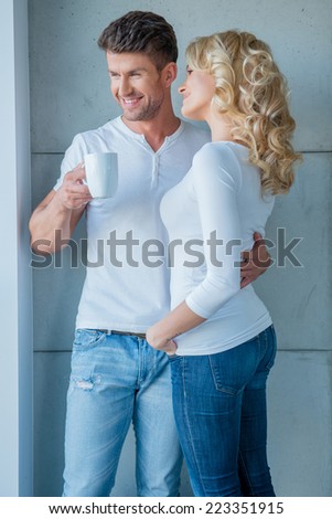 Affection couple standing looking out of a window smiling as they watch something outside while holding their morning coffee
