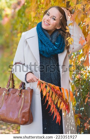 Stylish young woman in a polo neck and coat walking in a park picking decorative autumn leaves and looking at the camera with a lovely smile