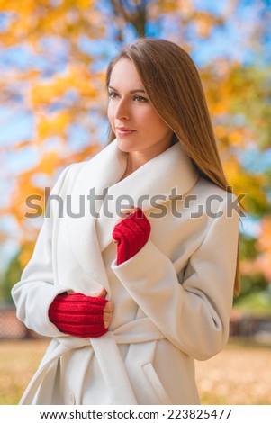 Elegant confident young woman in a stylish white overcoat and red mittens standing