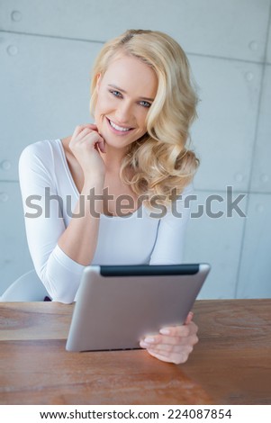 Happy woman with beautiful curly blond hair sitting at a table holding a tablet computer smiling at the camera