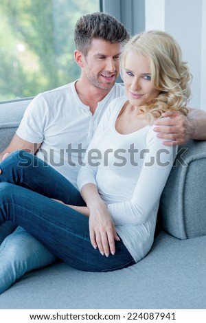 Romantic couple relaxing close together on a sofa in front of a window overlooking the garden looking at the camera with a friendly smile