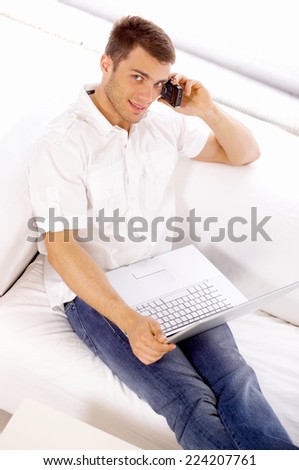 Handsome Young Salesman on White Couch Calling Someone Using Mobile Phone with Laptop on His Lap. Captured Indoor on White Background.