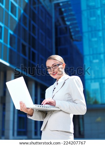 Pretty Young Businesswoman in White Posing with Laptop While Standing. Isolated on Blue Huge Building.