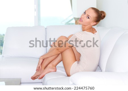 Sexy attractive slender blond woman sitting on a white sofa in front of a window in the living room looking back at the camera with parted lips