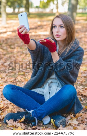 Smiling woman taking a selfie in an autumn park while sitting cross-legged on the ground posing for her mobile phone