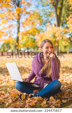 Happy Young Woman Sitting on Grassy Ground with Dry Leaves Using Laptop. Captured Outdoor During Autumn Season
