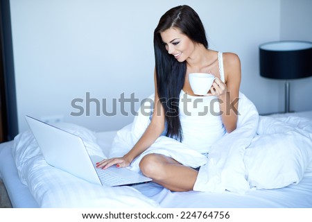Pretty Smiling Woman Busy Browsing on Laptop While Having a Cup of Coffee on White Bed. Captured Indoor.
