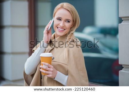 Attractive woman taking a call on her mobile phone smiling as she listens to the conversation with a mug of takeaway coffee in her hand