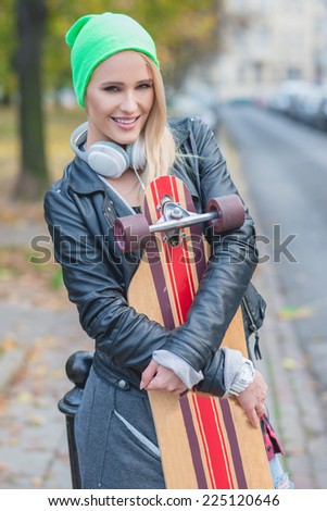 Smiling Young Pretty Woman in Trendy Attire Embracing Skateboard at Street Side  Looking at Camera.
