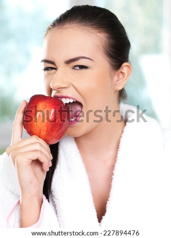 Close up Pretty Young Woman Wearing White Bathrobe After Shower Biting a Fresh Red Apple While Looking at the Camera.
