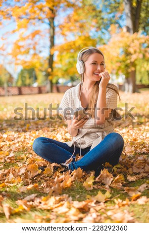 Natural young woman listening to music on headphones as she sits amongst the fallen autumn leaves in a park looking to the side with a happy smile