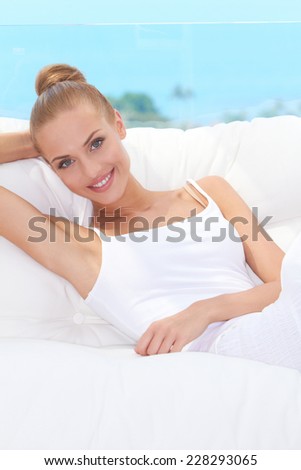 Smiling elegant woman relaxing on a white settee a patio with a blue sky background looking at the camera with a friendly smile