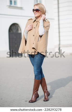 Elegant stylish attractive woman in a trendy outfit of boots and a cape standing in an urban square in her sunglasses
