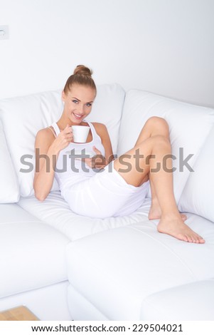 Pretty woman relaxing at home reclining bare foot on the sofa with a cup of coffee smiling at the camera