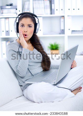 Close up Pretty Long Hair Woman in Surprise Face Expression  Wearing Casual Clothing and Sitting on White Couch with Crossed Legs While Listening Something From Laptop through Headphone.