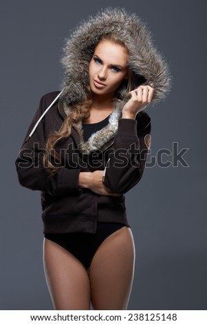 Pretty Sexy Young Woman in Black Swimsuit Covered with Gray Jacket with Furry Hood. Posing at the Studio with Gray Background While Looking at the Camera.