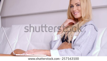 Close up Smiling Sexy Woman with Blond Hair  Wearing a Gray Sleep Robe  Sitting on a White Couch with Laptop on her Lap While Looking at the Camera.
