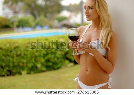 Waist Up of Blond Woman Wearing White Bikini Leaning Against Pillar and Holding Glass of Red Wine While Looking Contentedly into the Distance
