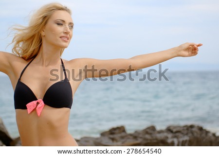Blond Woman Wearing Black Bikini with Pink Bow Standing with Arms Outstretched and Looking Blissfully into the Distance at Rocky Beach Shore