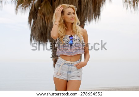 Three Quarter Length Image of Blond Woman Wearing Denim Cut Offs and Crop Top Brushing Hair from Face and Standing on Beach with Palm Trees