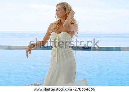 Smiling Blond Woman Wearing Strapless Sun Dress Leaning on Railing with Hands in Hair Standing on Ocean Front Resort Glass Balcony