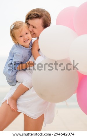 Loving mother and her small daughter share a tender happy moment as she holds the toddler in her arms with a bunch of pink and white party balloons.