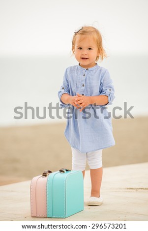 Adorable little girl standing outdoors at the seaside with two small suitcases in front of her smiling at the camera