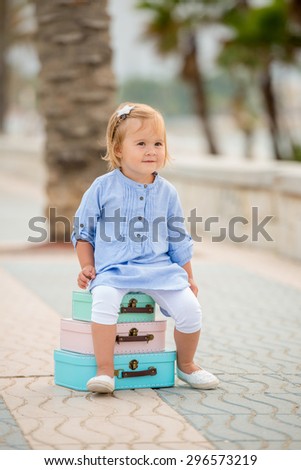 Adorable little girl on a summer vacation sitting on a stack of three small colorful suitcases on a tropical sidewalk with palm trees
