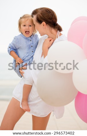 Happy young mother with her small daughter in her arms nuzzling her forehead with her face in a laughing fun gesture as they stand outside with pink and white party balloons.
