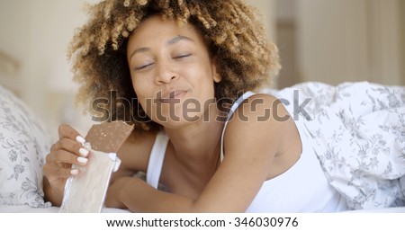 Cute young african-american woman eating chocolate in bed at home