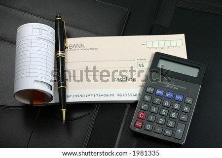 An open cheque book, calculator, and a black-gold roller ball pen lying on top of a black executive leather folder.