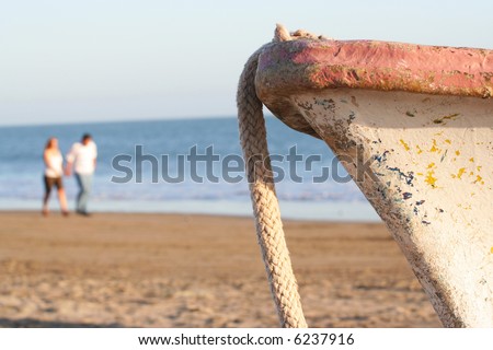 A blurred couple in the background walk past the bow of a fishing boat in coastal Mexico.