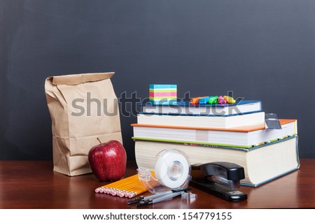School books with apple and  paper bag lunch on desk in front of a blank chalkboard.
