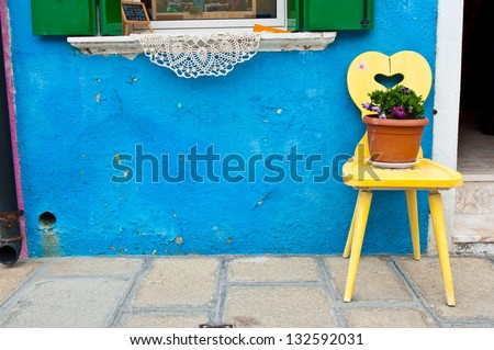 A yellow wooden chair placing in front of a bright blue house on Burano island, Italy