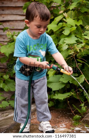 content young child watering the garden with a green colored hose.  He is wearing a blue coolred to grey tracksuit tracksuit. In the background there vine leaves growing up against a wooden fence