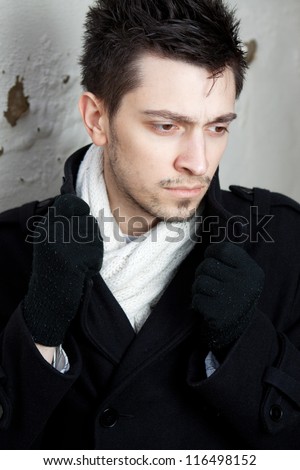 Handsome young man wearing a white scarf dark colored jacket and gloves. Standing against a light colored old wall.  He has brown eyes, and brown hair, and facial hair that is more than just stubble