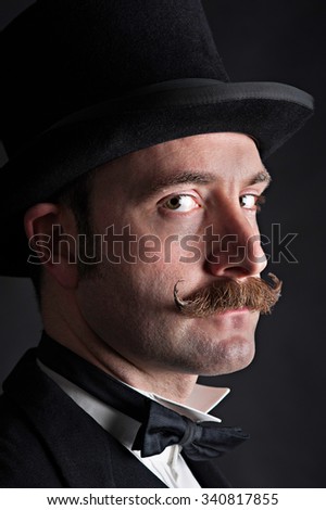 Low key portrait of male wearing a black top hat. He has a moustache and is looking sideways on into the camera. He has a straight expression on his face. He could be a magician or Victorian gent.