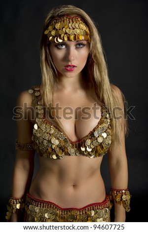 This is an image of a blonde haired and blue eyed belly dancer wearing red and gold coined outfit.  She has a seductive and mysterious expression on her face as she looks past the camera