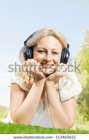 Happiness blonde woman with headphones relaxing on green grass in the park.
