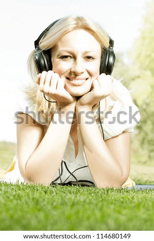 Happiness blonde woman with headphones relaxing on green grass in the park.